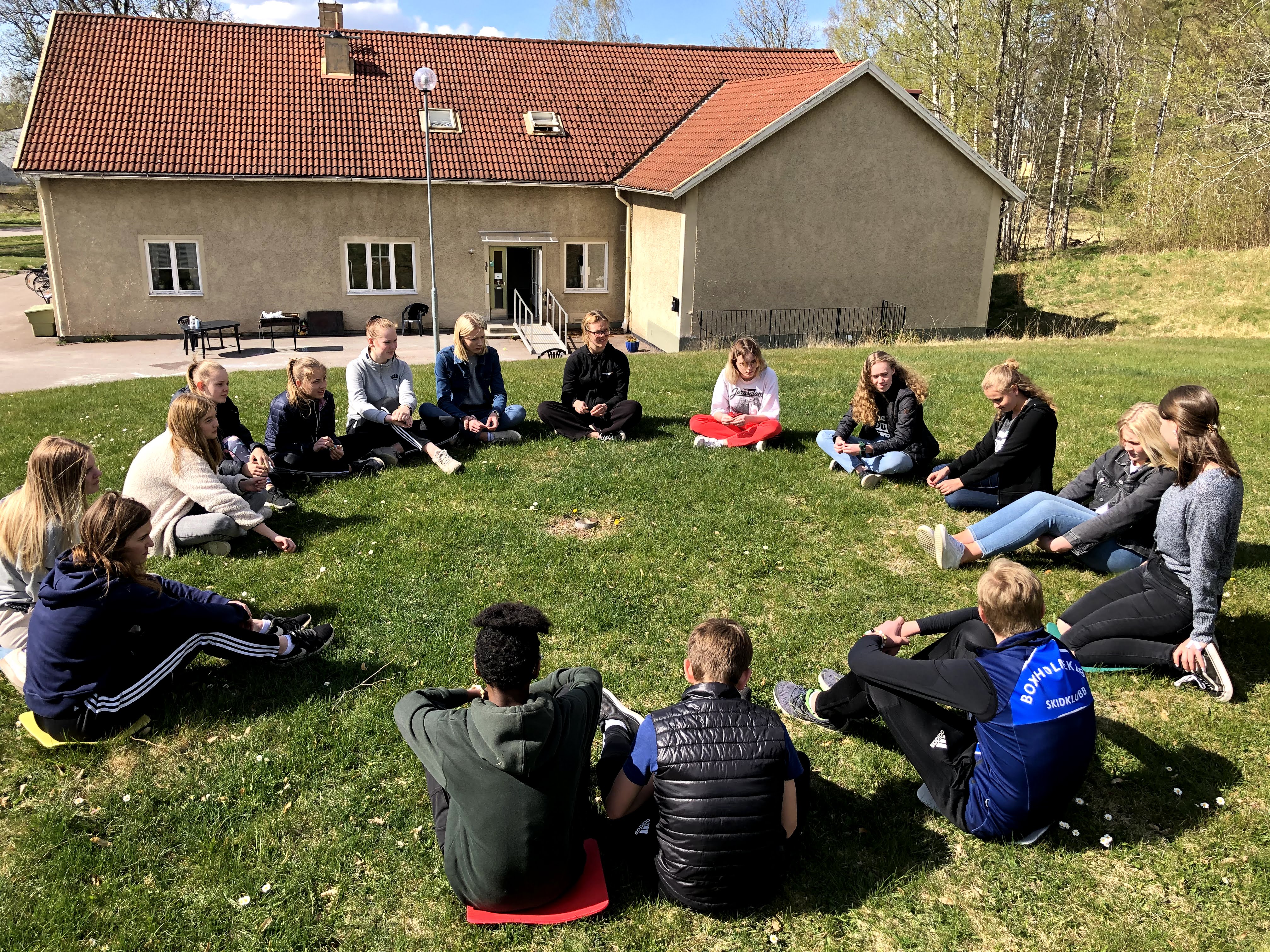 Ungdomar sitter i en ring på gräsmattan framför Boxholms församlingshem.