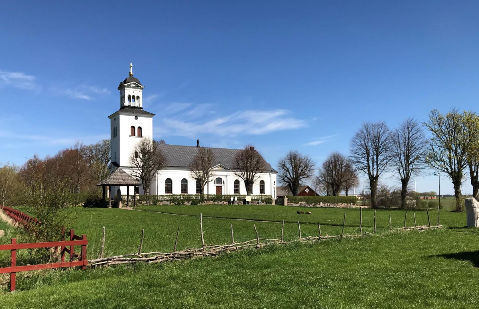 Röks kyrka. En vit stor kyrka med torn och öppen lanternin. Framför kyrkan står Rökstenen, en runsten under ett tak vid kyrkmuren. I förgrunden ett gärdsgård och ett rött staket.