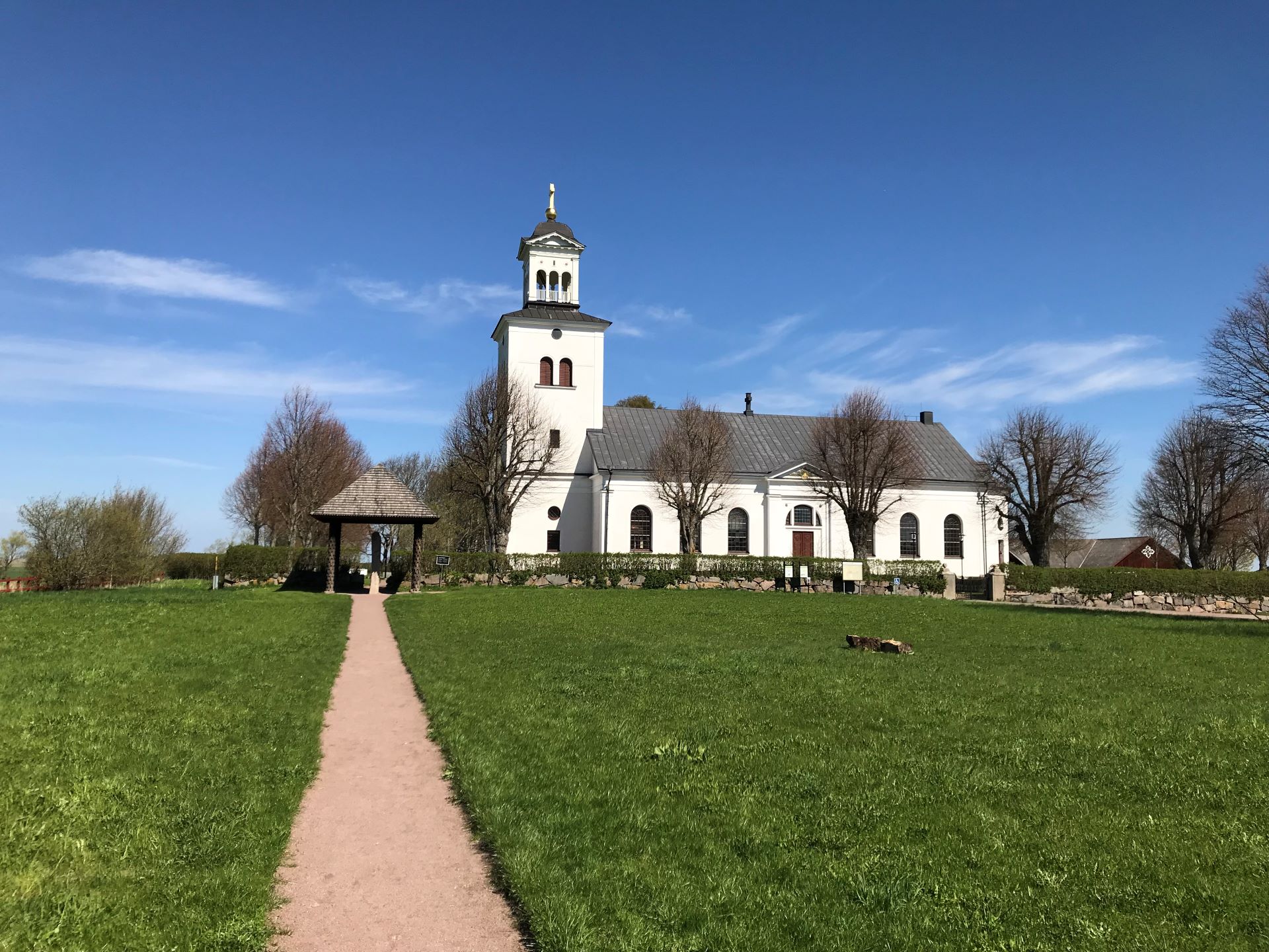 Röks kyrka. En vit kyrka med plåttak. En grusgång går rakt fram till Rökstenen, en runsten som står under ett tak vid kyrkmuren.