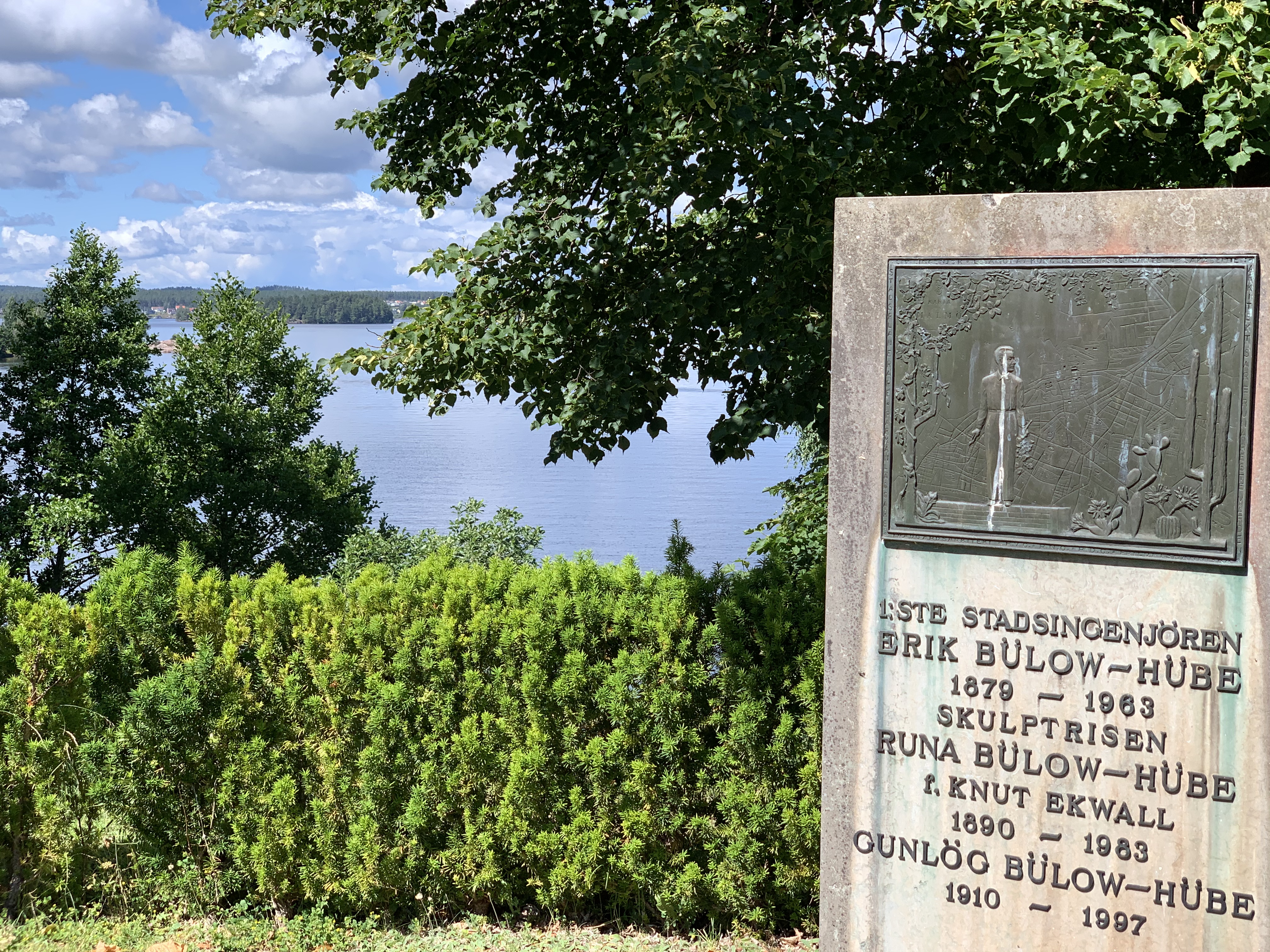 Gravsten på Blåviks kyrkogård. Utsikt över sjön Sommen. Träd och en häck i förgrunden.