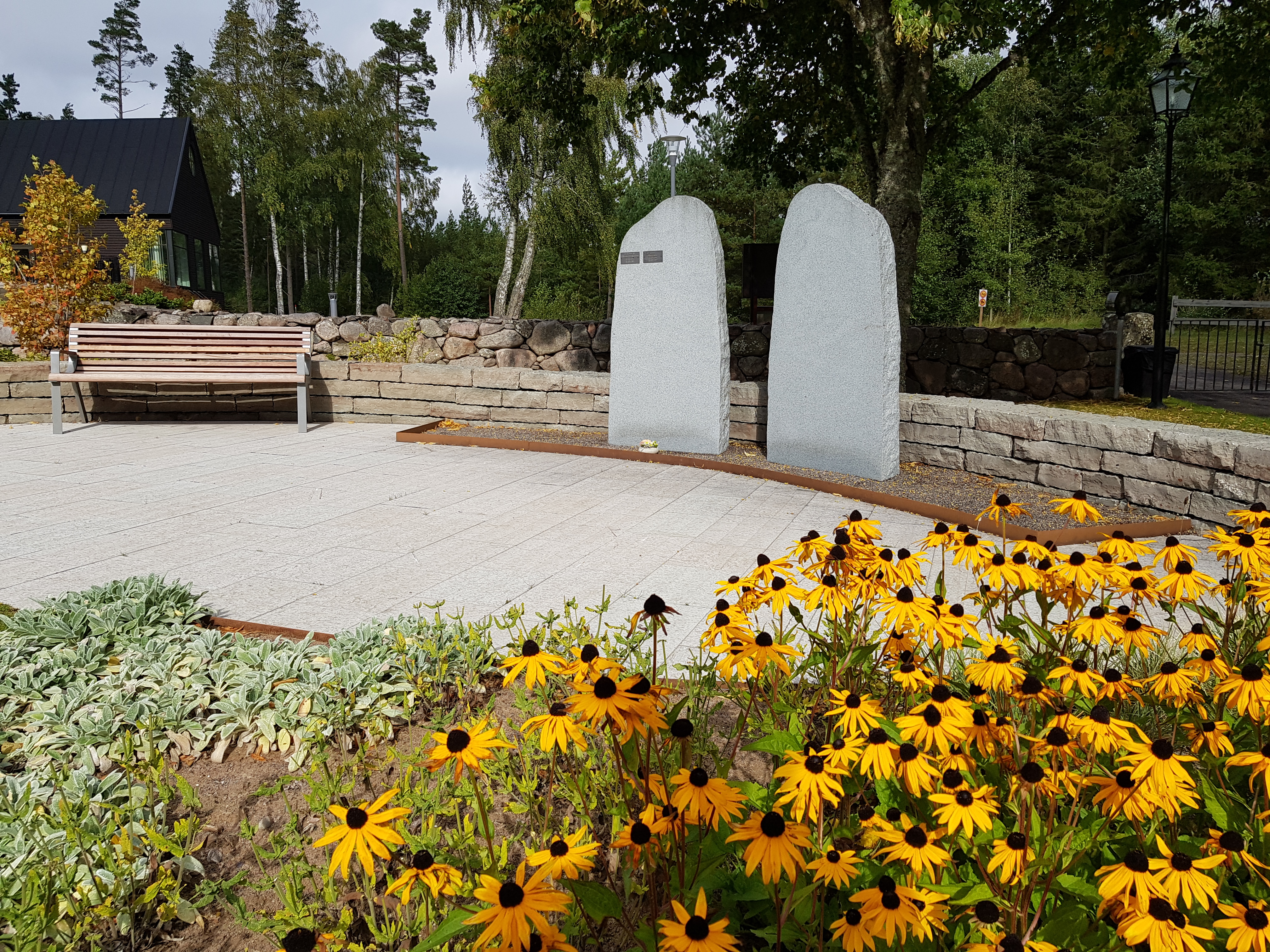 Två stenar med namnplattor. En soffa vid sidan om och gula blommor i förgrunden.