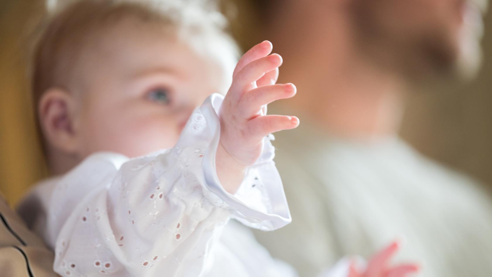 En bebis klädd i dopdräkt sträcker fram sina händer.  