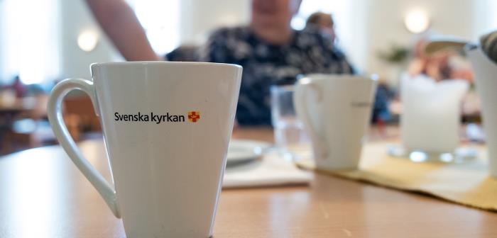 En vit kaffekopp med Svenska kyrkans logotyp står på ett bord. I bakgrunden syns människor som deltar i kyrkans verksamhet.