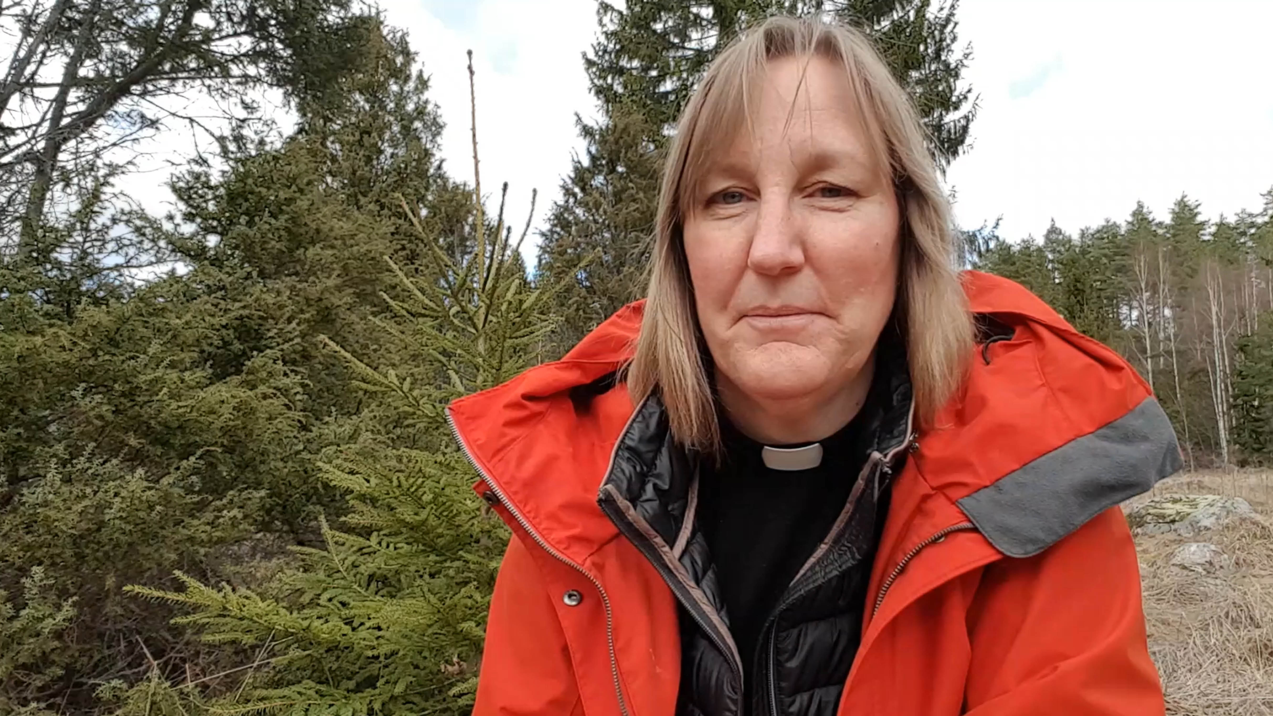En kvinnlig präst med halvlångt, blont hår, röd jacka, svart skjorta och prästkrage står i en skog. I bakgrunden syns gröna granar.