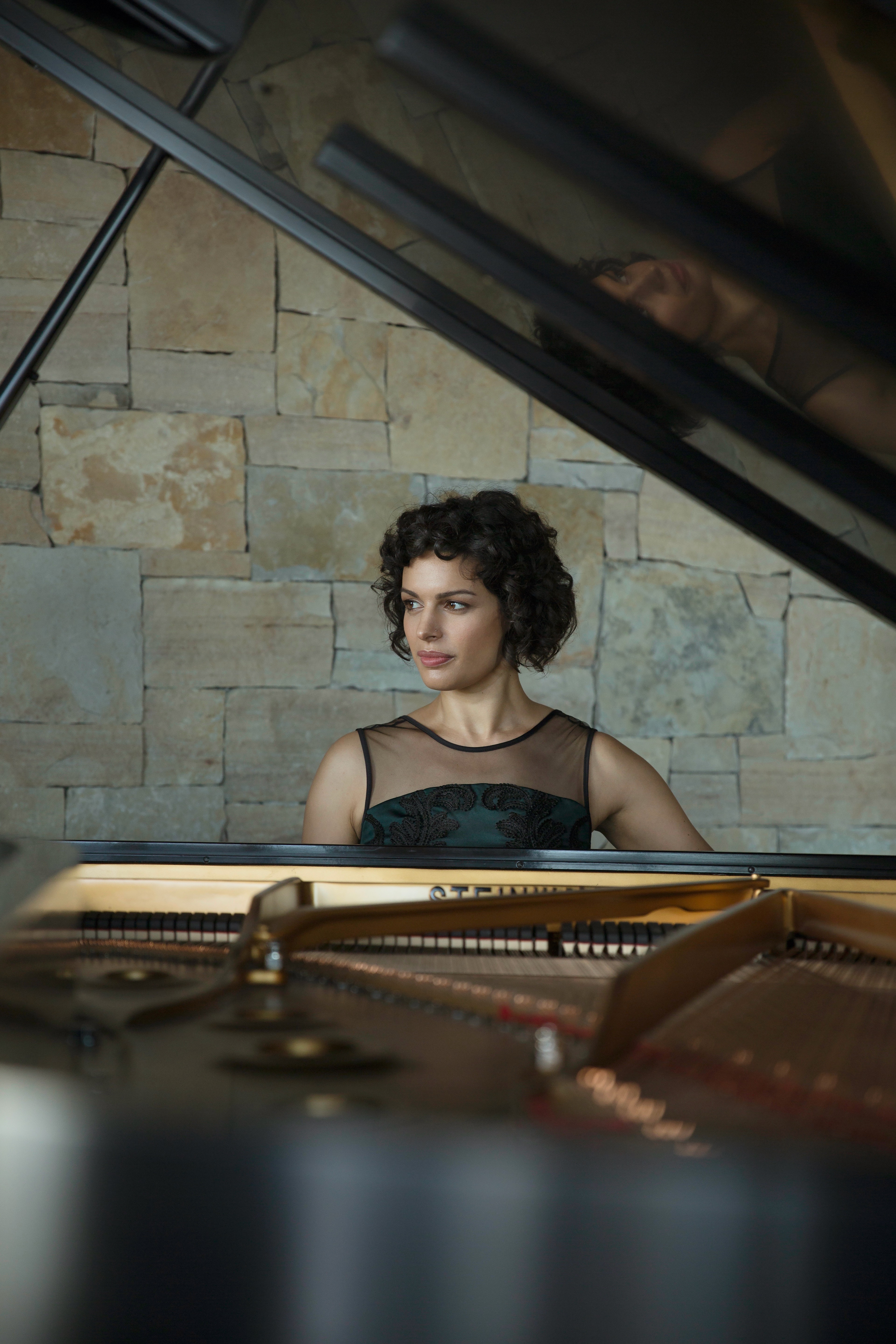 En kvinna med svart klänning sitter vid en flygel och ser ut över sin högra axel.