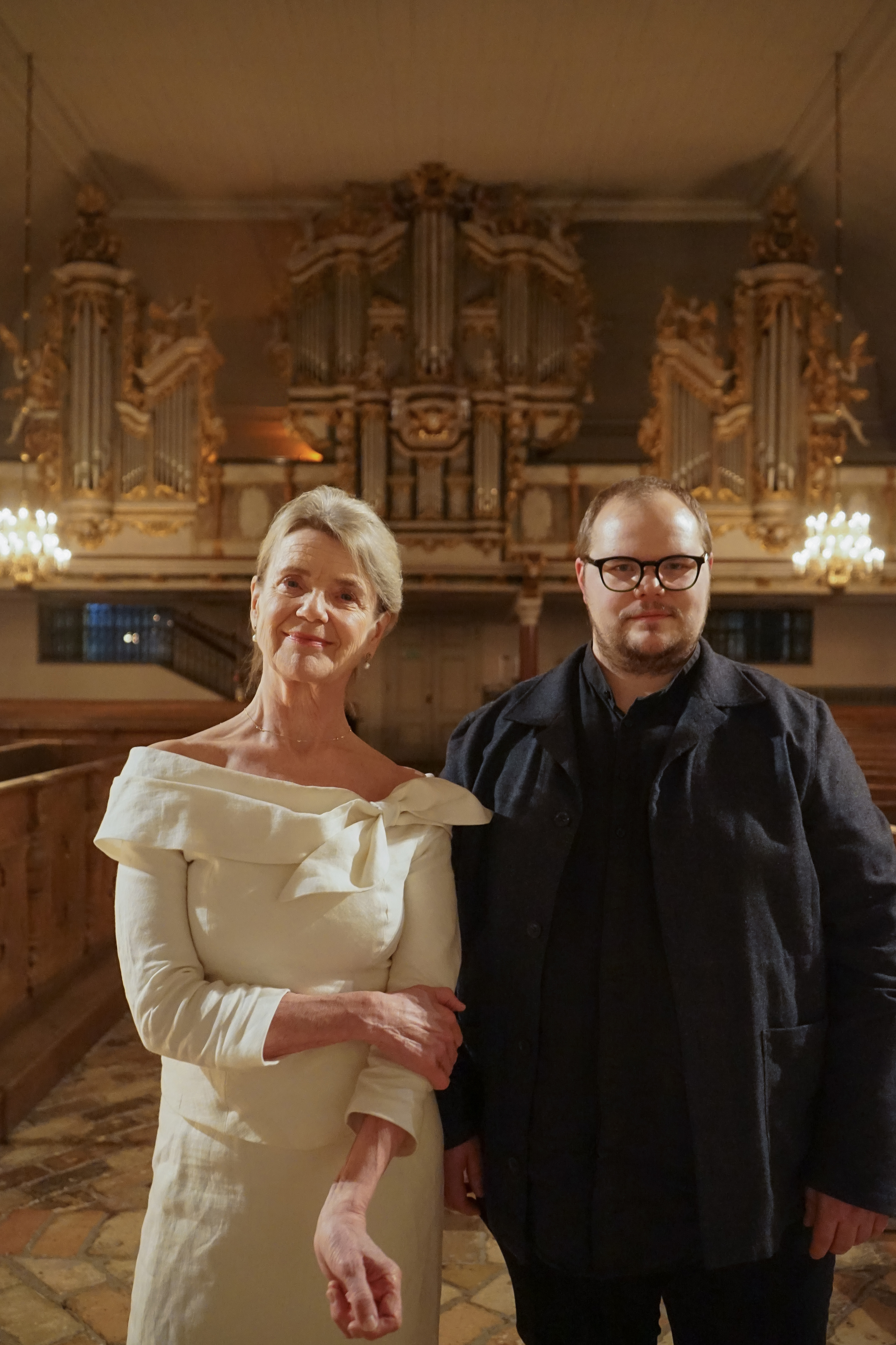 En kvinna med vit klänning och blont/grått kort hår, står bredvid en man med svarta kläder, glasögon och brunt kort hår. De står i Lövstabruks kyrka med Cahmanorgeln bakom dem.