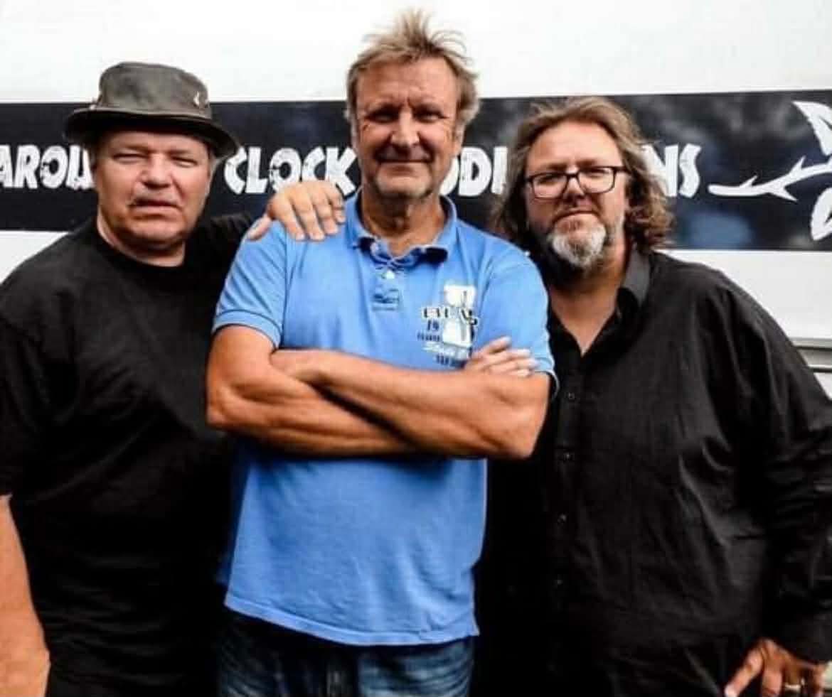 Tre män i en bandkonstellation står på rad och ser in mot kamrean.