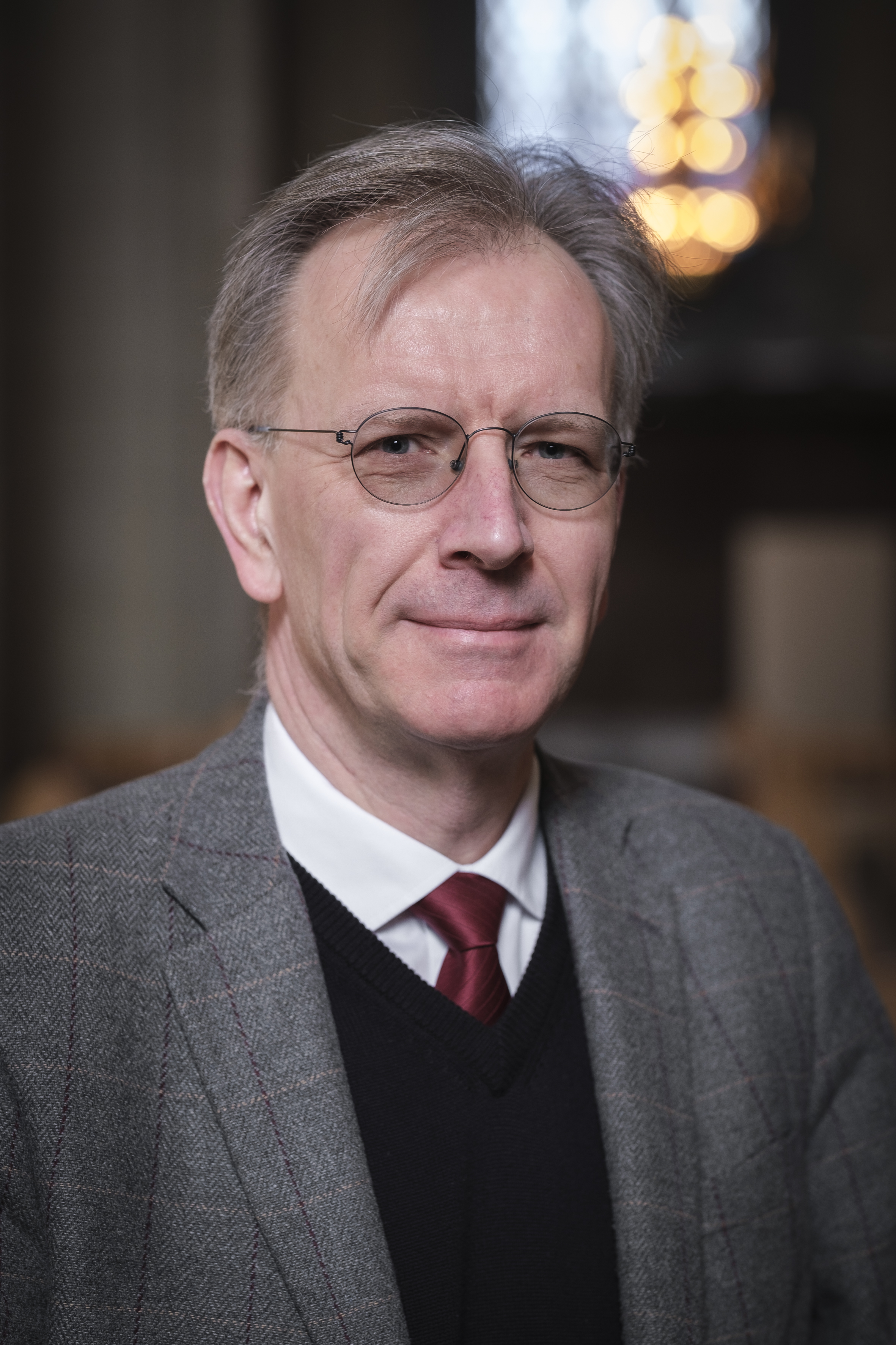 Ulric Andersson, svensk kyrkomusiker och operasångare, står klädd i kostym och ler försiktigt mot kameran.