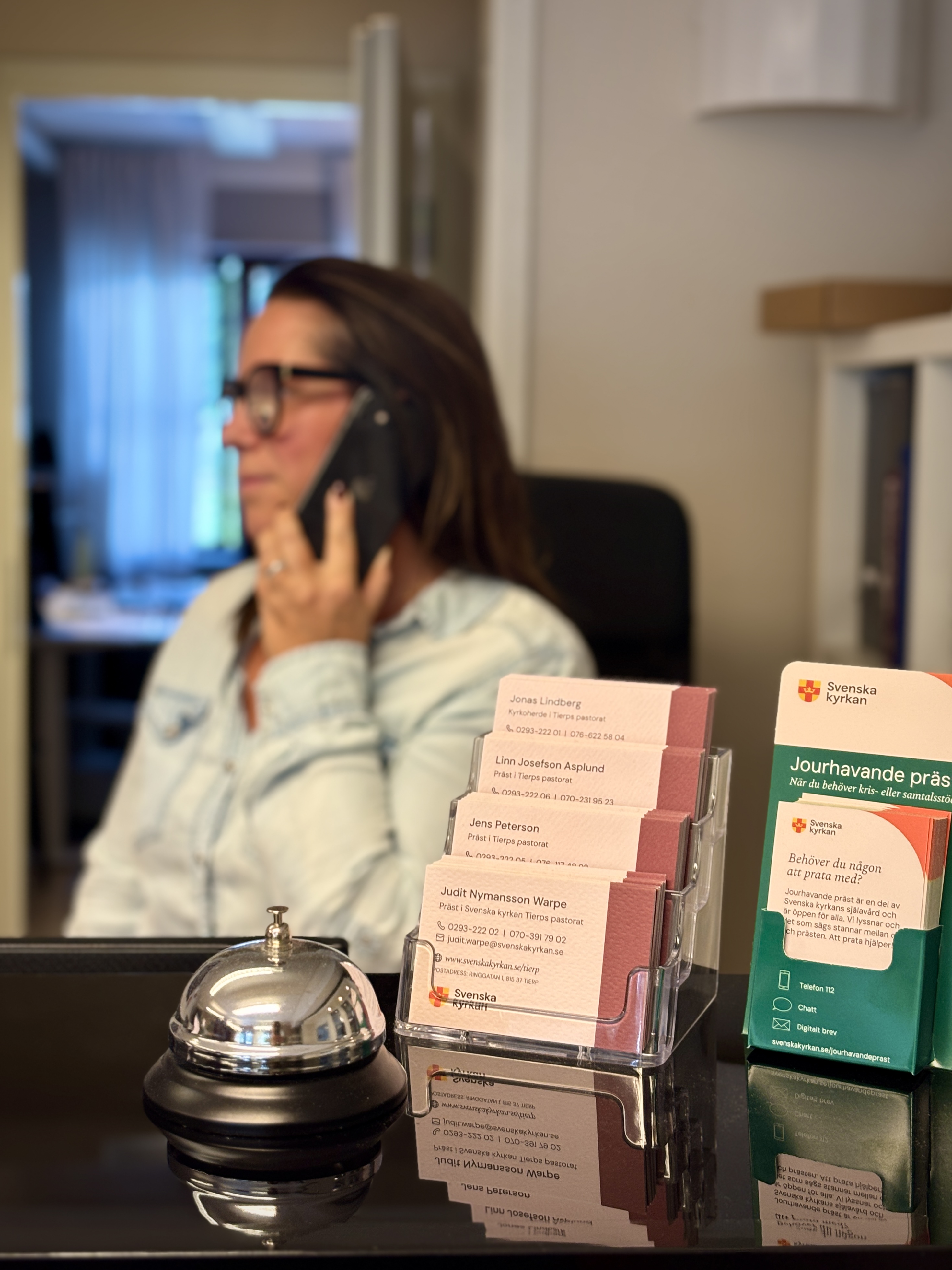 En kvinnlig receptionist sitter vid en expedition och tar emot samtal, i förgrunden står visitkort med kontaktuppgifter till anställda präster.
