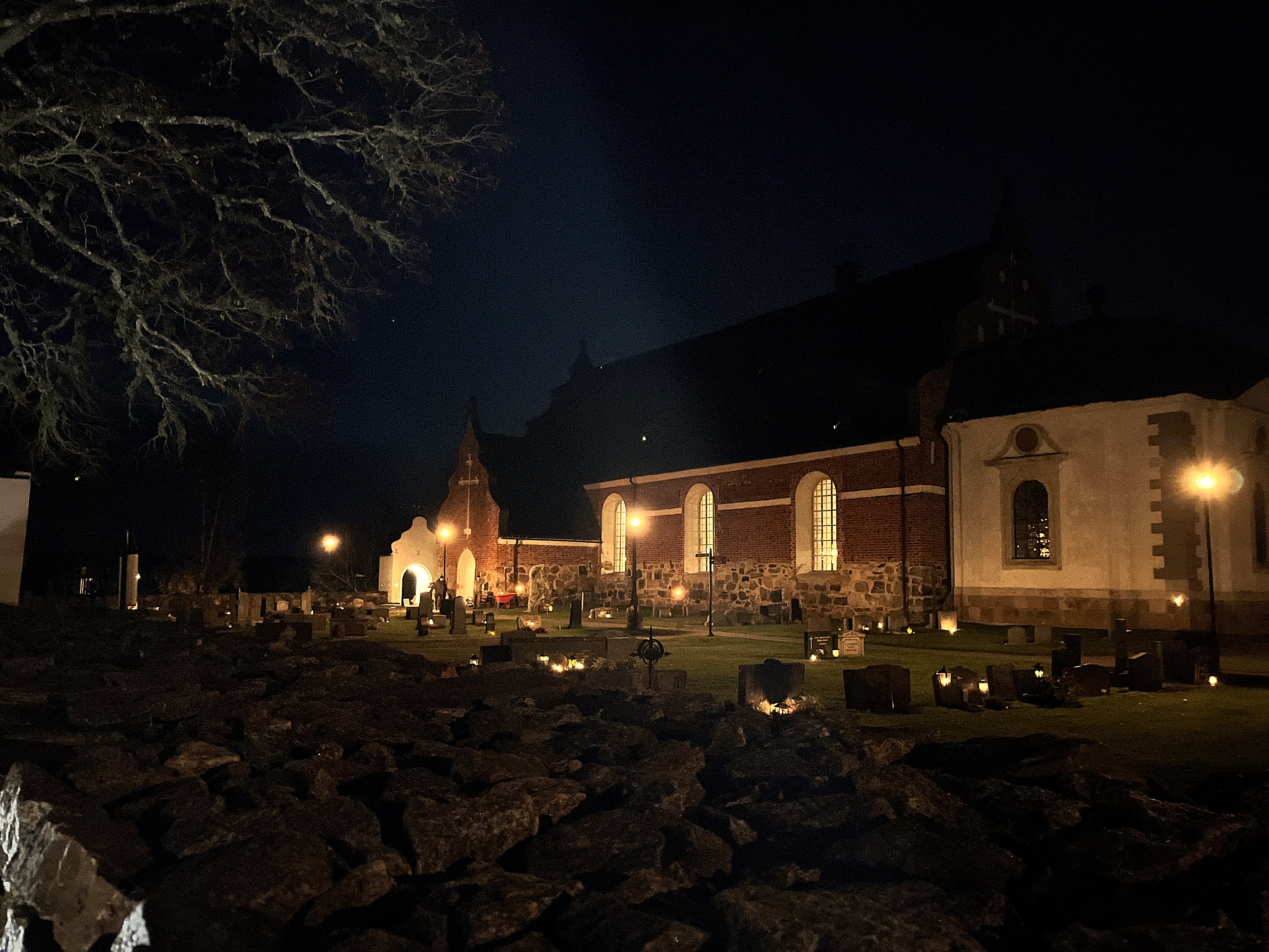 Österlövsta kyrka i allhelgonatid. Förgrund på kyrkogården med tända ljus i mörkret, Österlövsta kyrka i bakgrunden.