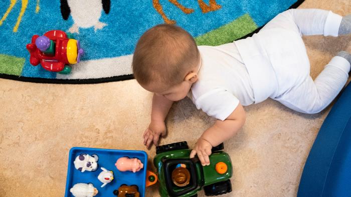 Bilden föreställer en bebis på golvet som leker med en traktor med släp. 