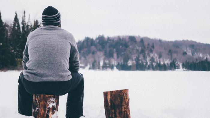 En man sitter på en stubbe och blickar ut över en snötäckt sjö.