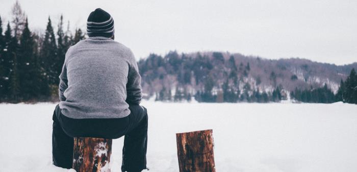 En man sitter på en stubbe och blickar ut över en snötäckt sjö.
