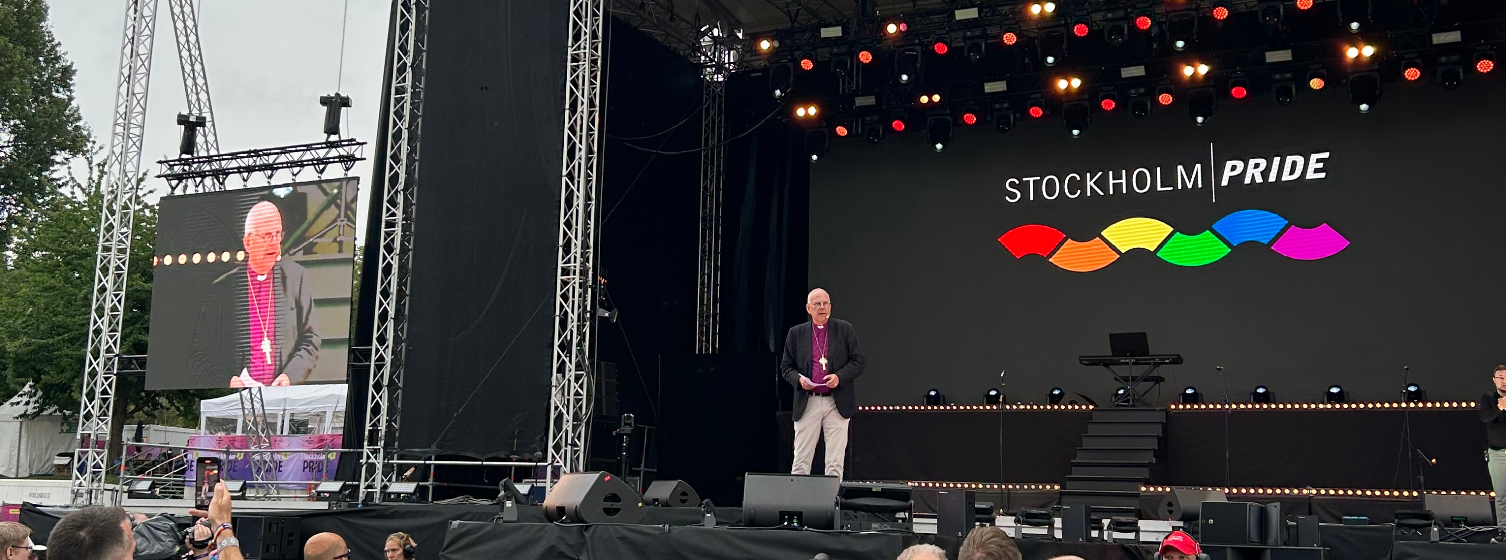 Ärkebiskop Martin Modéus står på en scen och håller tal. Bredvid scenen visas han på en storbilds-tv.  