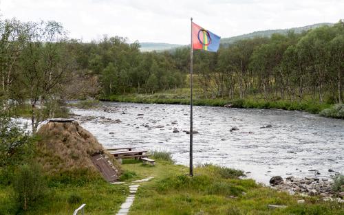 Samisk flagga i flaggstång vid ett vattendrag.