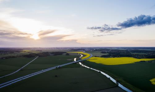 Flygfoto där ett sommarlandskap, med åkrar och vägar, möter himlen, med en sol som lyser igenom molnen.