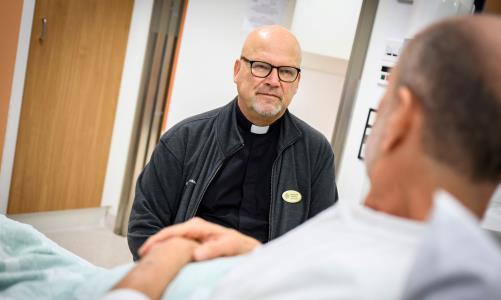 En man med prästkrage sitter hos en man som ligger i en sjukhussäng.