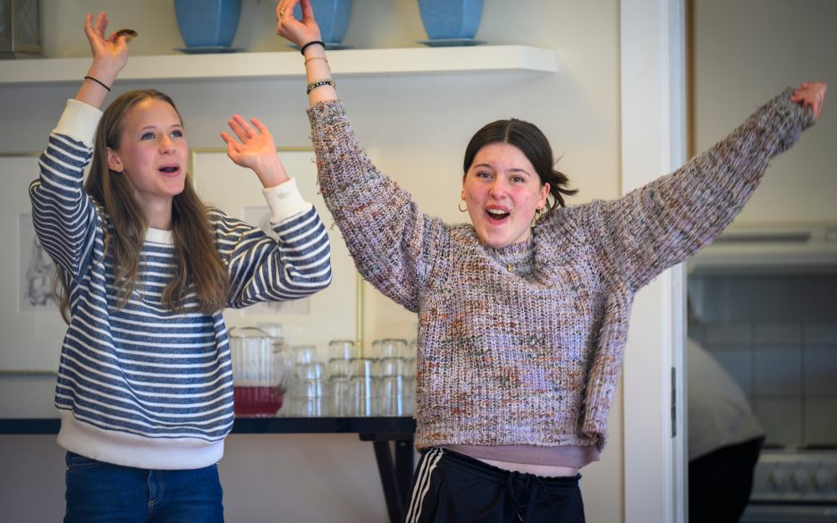 Två unga tjejer dansar glatt och viftar med armarna inomhus.