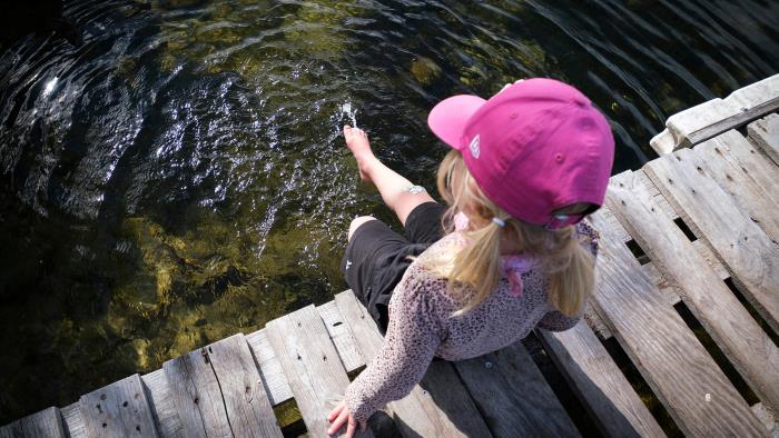 En liten flicka med rosa keps sitter på en brygga och doppar i tårna i vattnet.