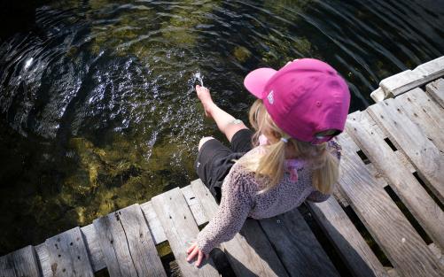 En liten flicka med rosa keps sitter på en brygga och doppar i tårna i vattnet.