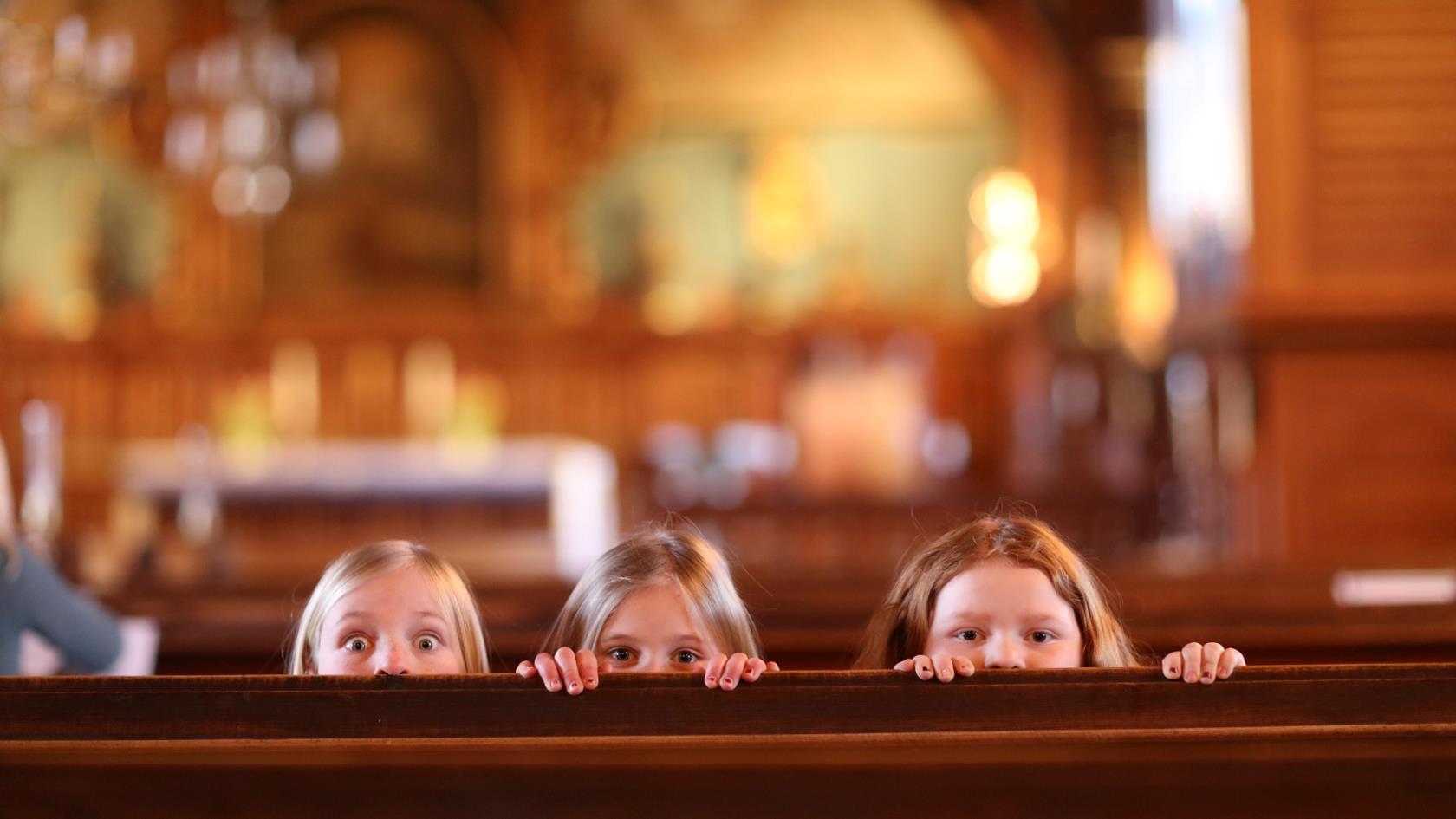 Tre flickor tittar upp bakom en kyrkbänk.