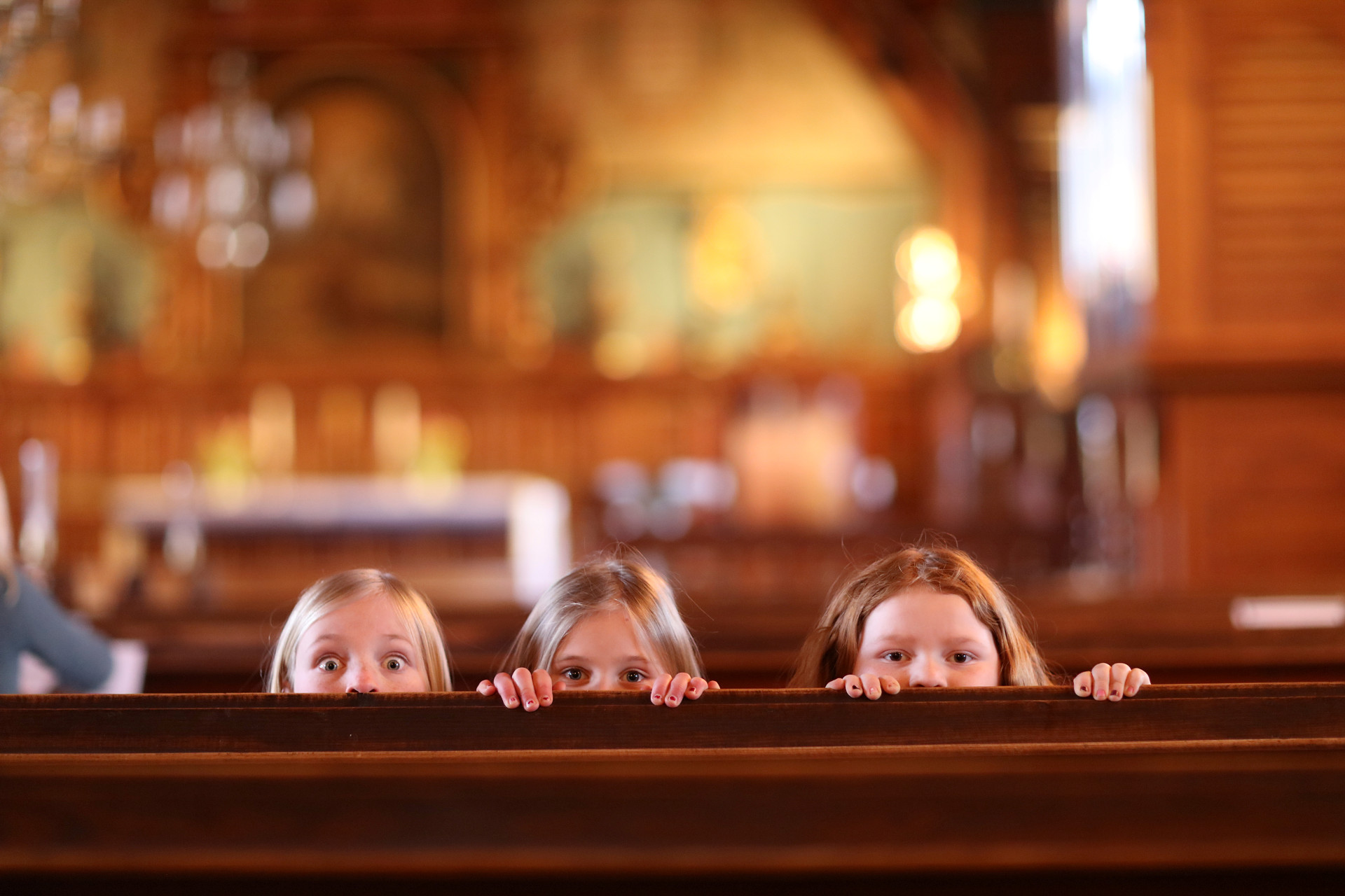 Tre flickor tittar upp bakom en kyrkbänk.