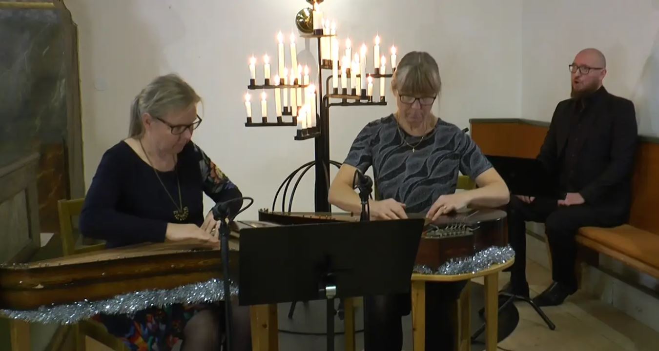 Två kvinnor sitter och spelar kantele. Man sjunger i bakgrunden.