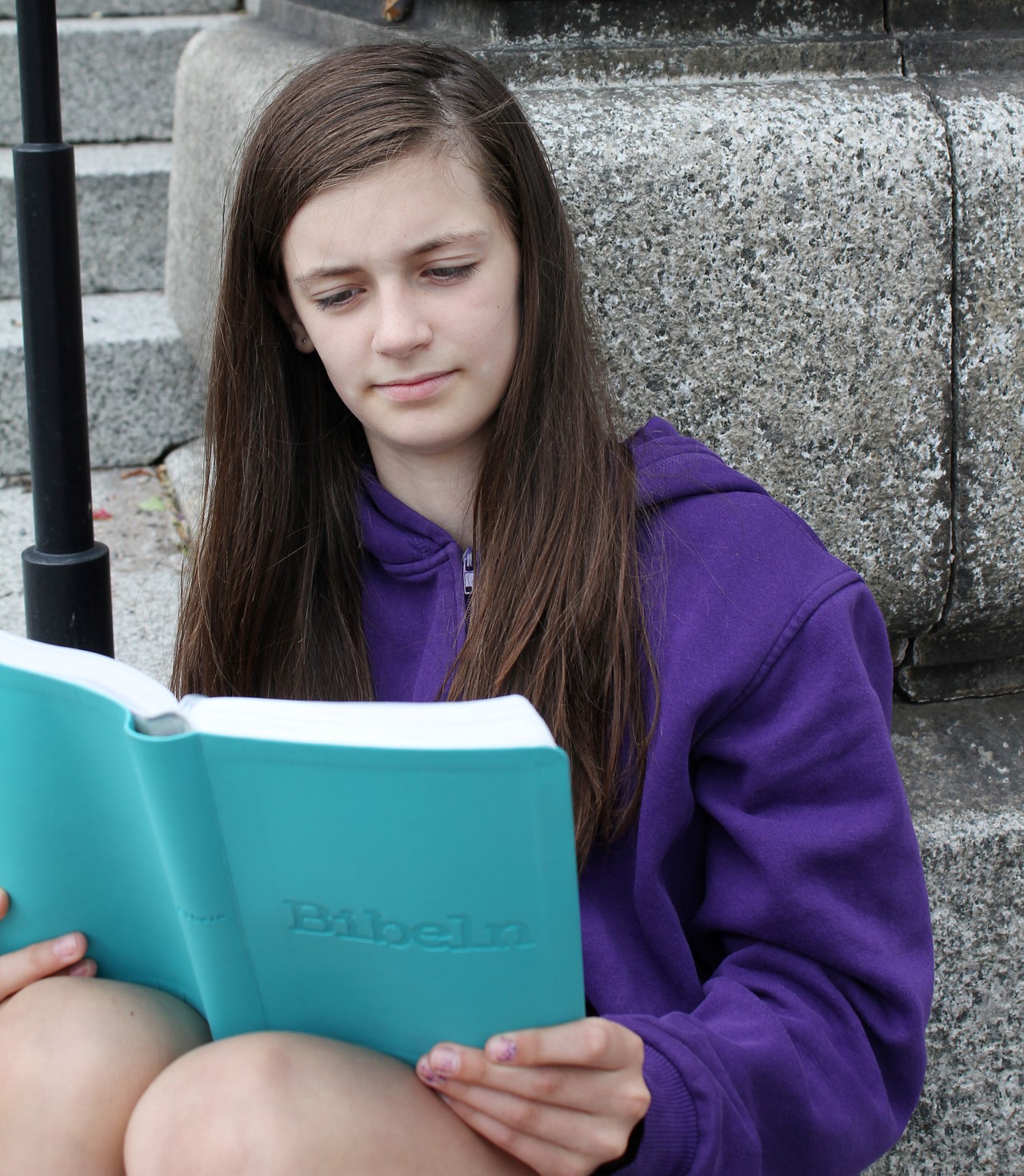 En tonårsflicka sitter lutad mot en stenmur och läser ur en turkos bibel.
