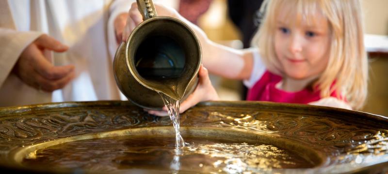 En liten flicka häller upp vatten från en kanna i en dopfunt.