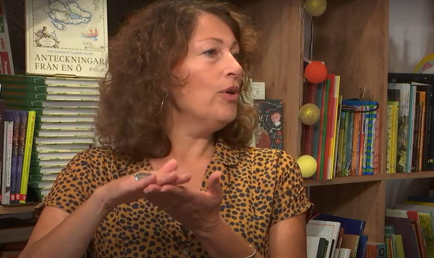Kvinna sitter framför en bokhylla, pratar och gestikulerar.