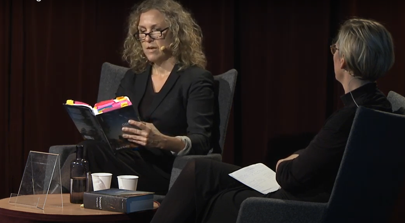 Två kvinnor på en scen. Den ena läser ur en bok.