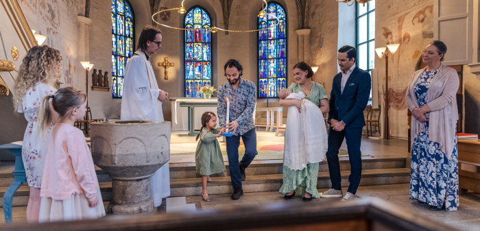 En glad dopfamilj, faddrar och präst står långt fram i kyrkan.