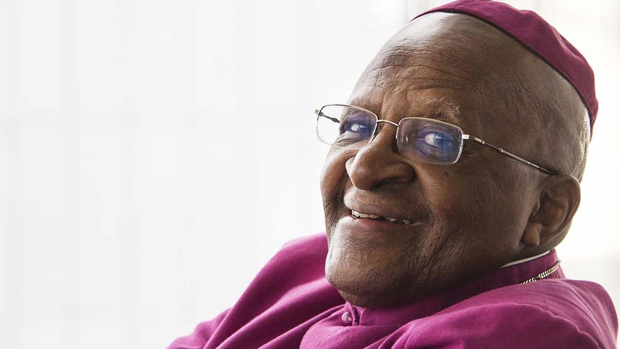 Leende Desmond Tutu i lila skjorta, med armarna i kors.