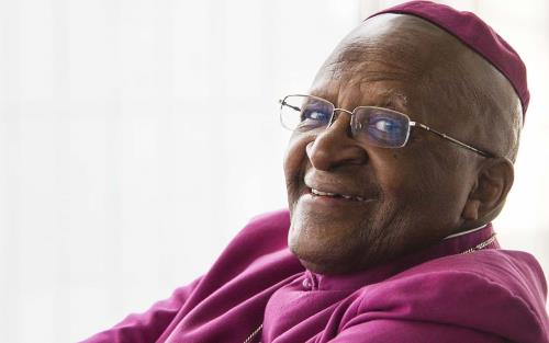 Leende Desmond Tutu i lila skjorta, med armarna i kors.