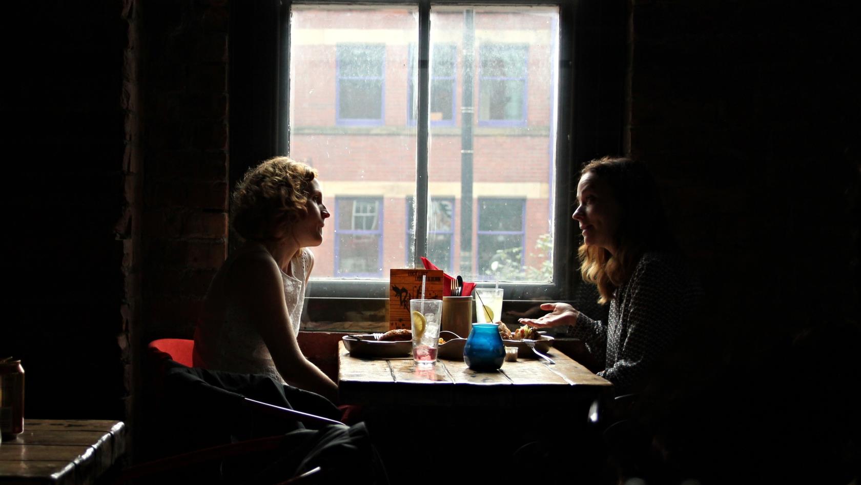 Två kvinnor sitter vid ett bord vid ett fönster och samtalar.