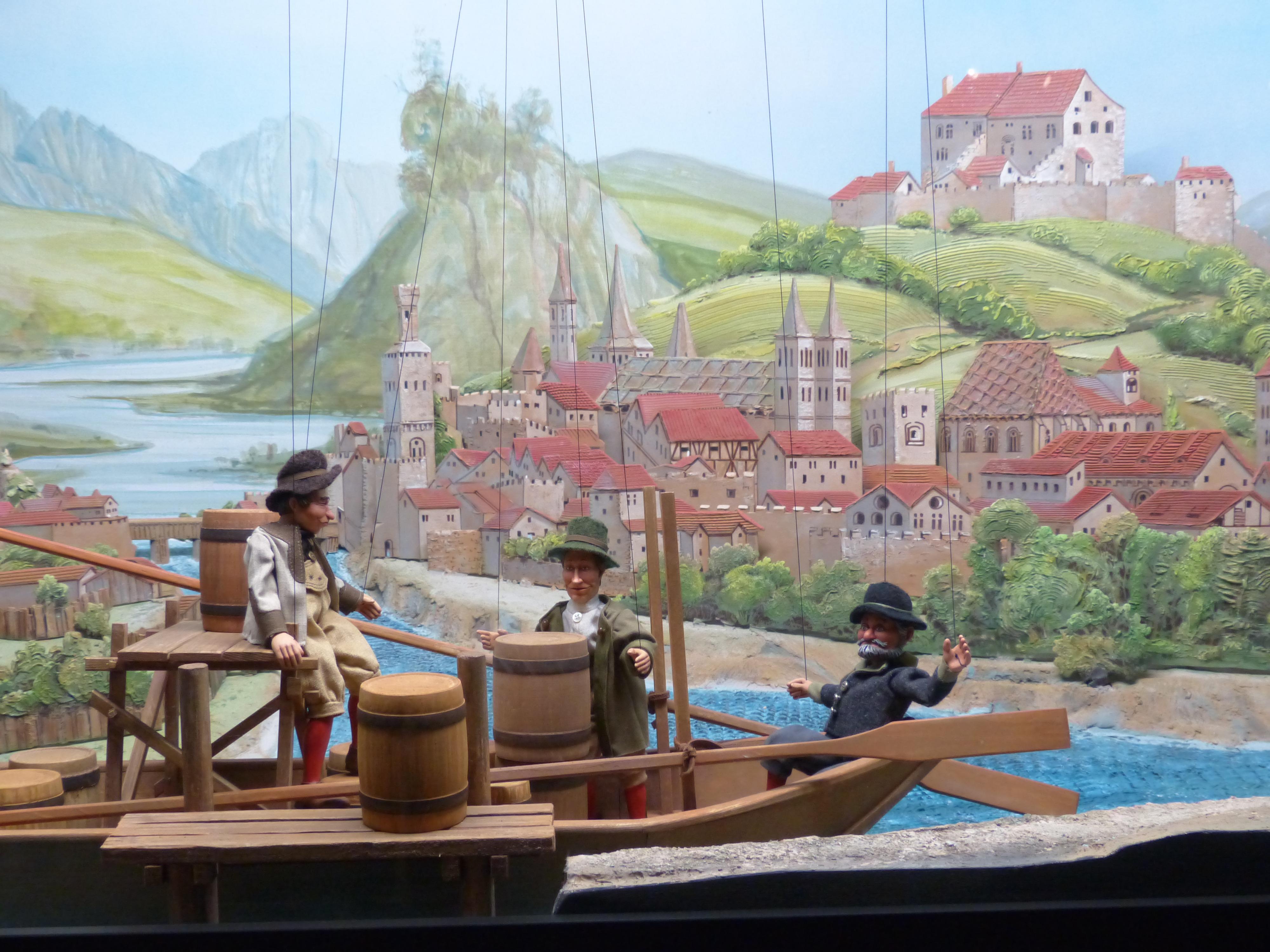 Marionettdockor vid en modellbåt. I bakgrunden en målad bild av en stad.