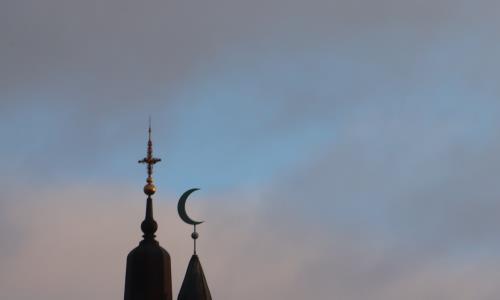 En silhuett av en kyrkas korskrönta torn och månen på en moske.