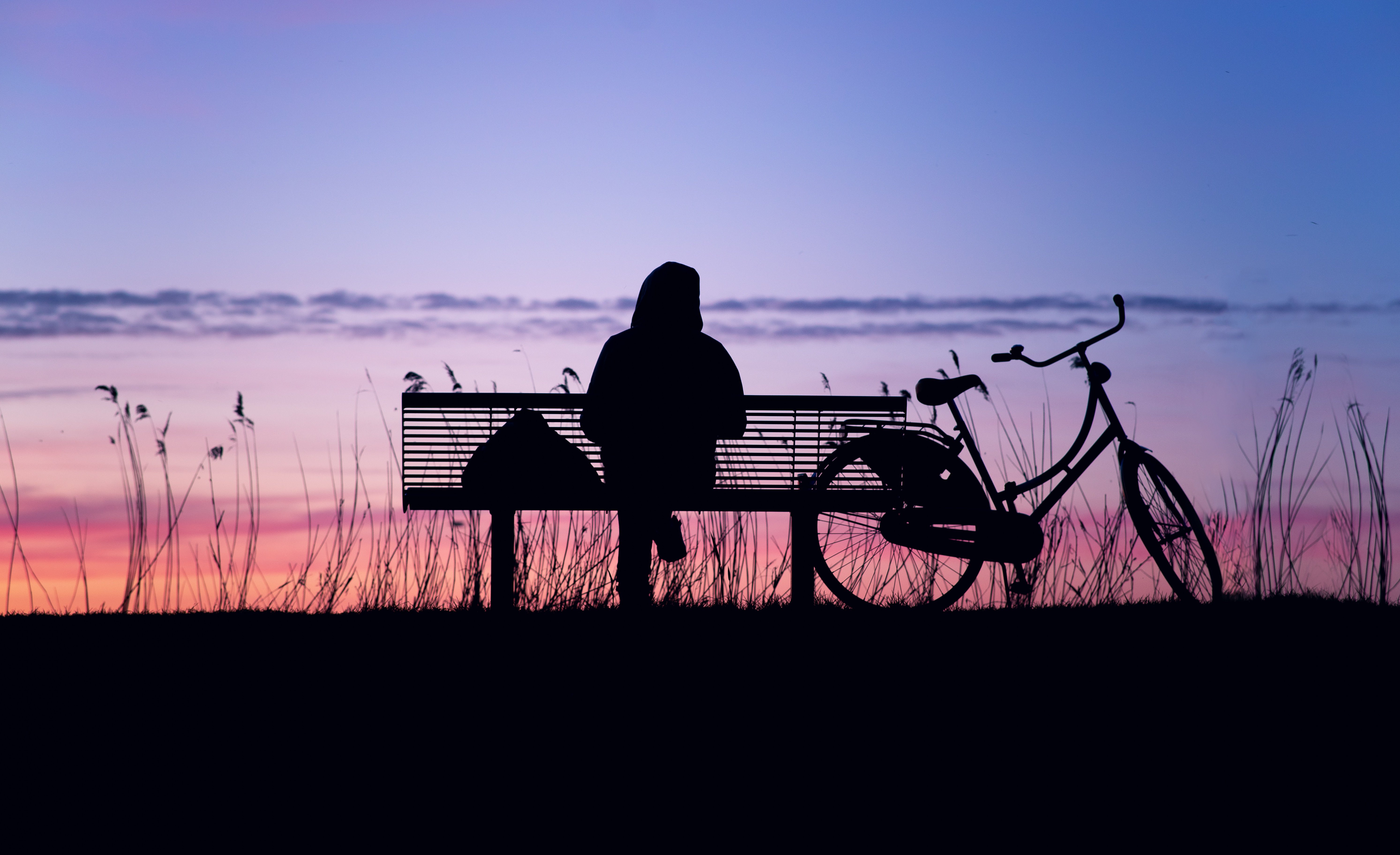 Silhuett av en person sittande på en parksoffa med en cykel bredvid. Himlen är blå och rosa.