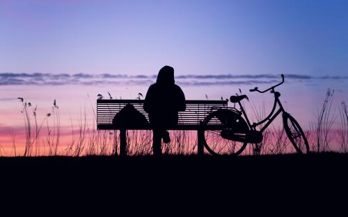 Silhuett av en person sittande på en parksoffa med en cykel bredvid. Himlen är blå och rosa.