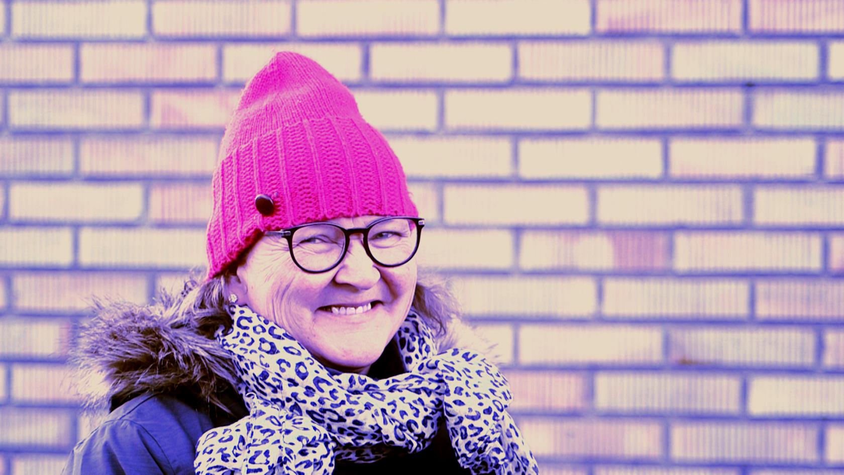 Tuula Polojärvi står i rosa mössa framför en tegelvägg och ler in i kameran.