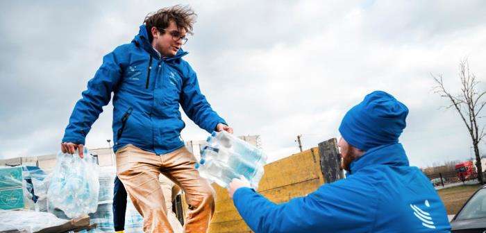 Två män i blå hjälporganisationsjackor lastar av vattenflaskor från ett lastbilsflak.