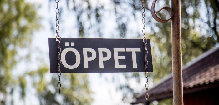 Skylt med texten öppet. I bakgrunden trädgrenar mot himmel.