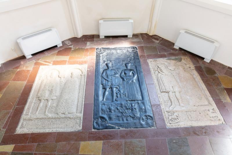 Tre gravstenar i bredd i golvet i koromgången. De yttersta gravstenarna är i ljus sten och den i mitten är svart. Stenarna visar avbildningar av vem som ligger begravda där. 