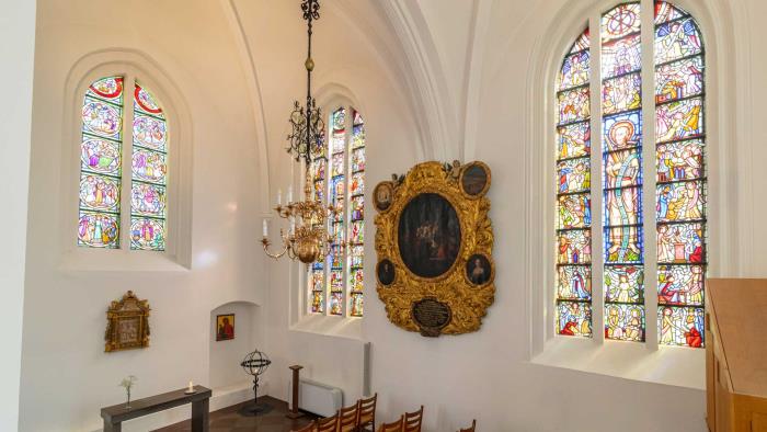 Vitkalkat kapell med höga målade glasfönster, lösa stolar, ljuskrona, ljusbärare, ikonmålning och altare. 