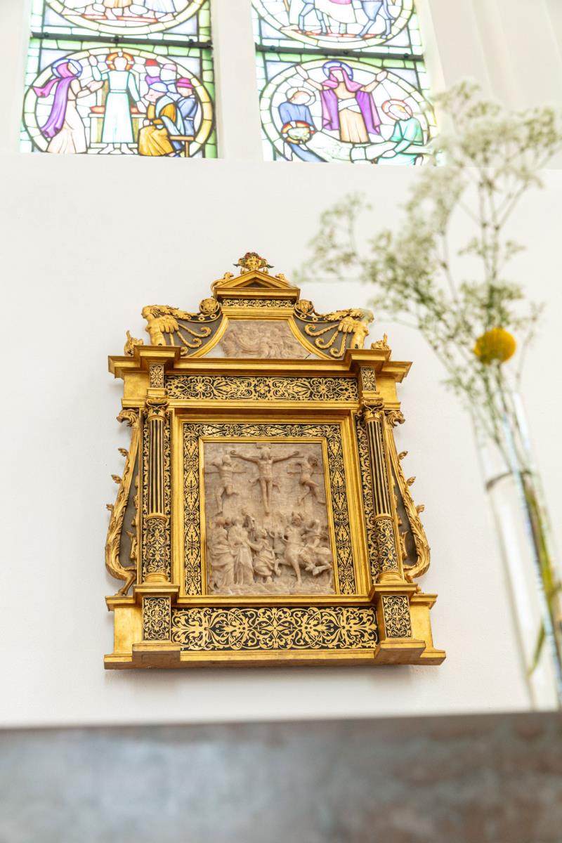 Rikt dekorerad guldram med en mittavla i sten. Hänger på en vitkalkad kyrkvägg nedanför ett målat glasfönster. I förgrunden en blomkvist och en altarskiva i sten.