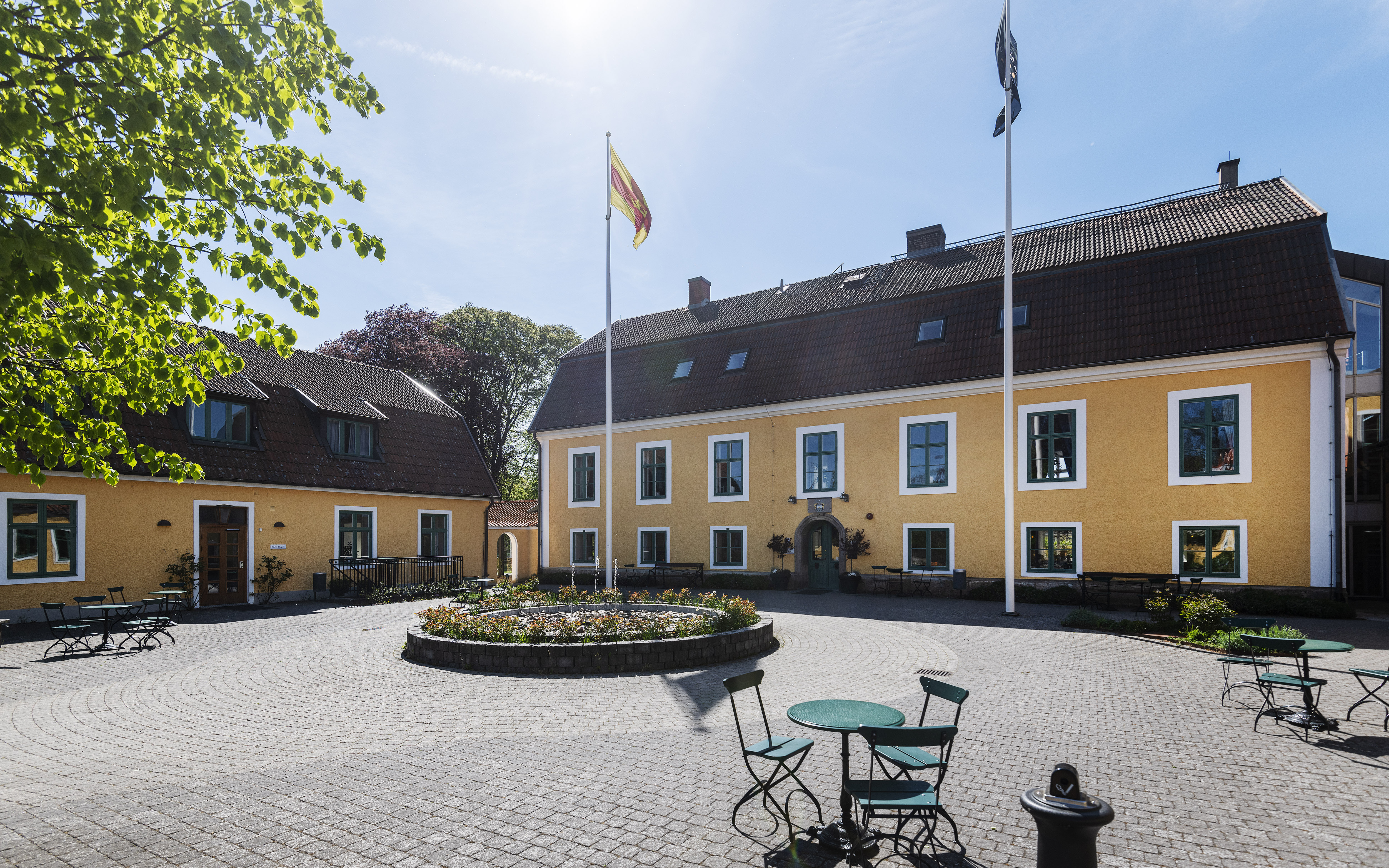 Gul herrgård med stor stenlagd innergård. Himlen är blå och Svenska kyrkans och svenska flaggan vajar i varsin flaggstång.