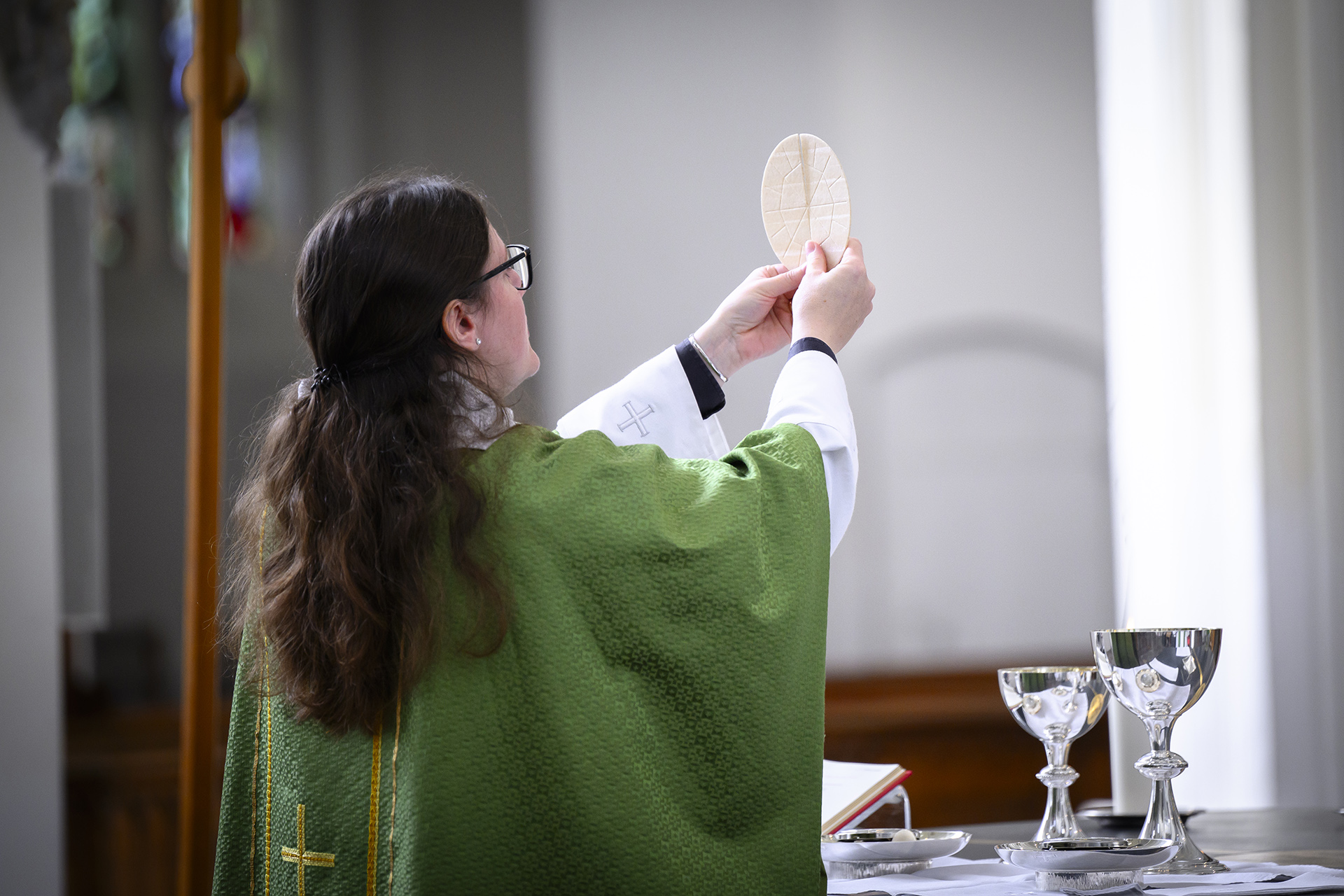 Kvinnlig präst med långt mörkt hår klädd i prästkappa lyfter bröd med händerna