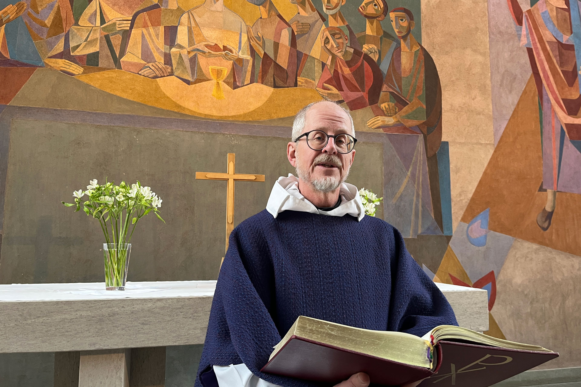 Äldre man med skägg och glasögon i liturgiska kläder med stor Bibel i händerna framför altare.