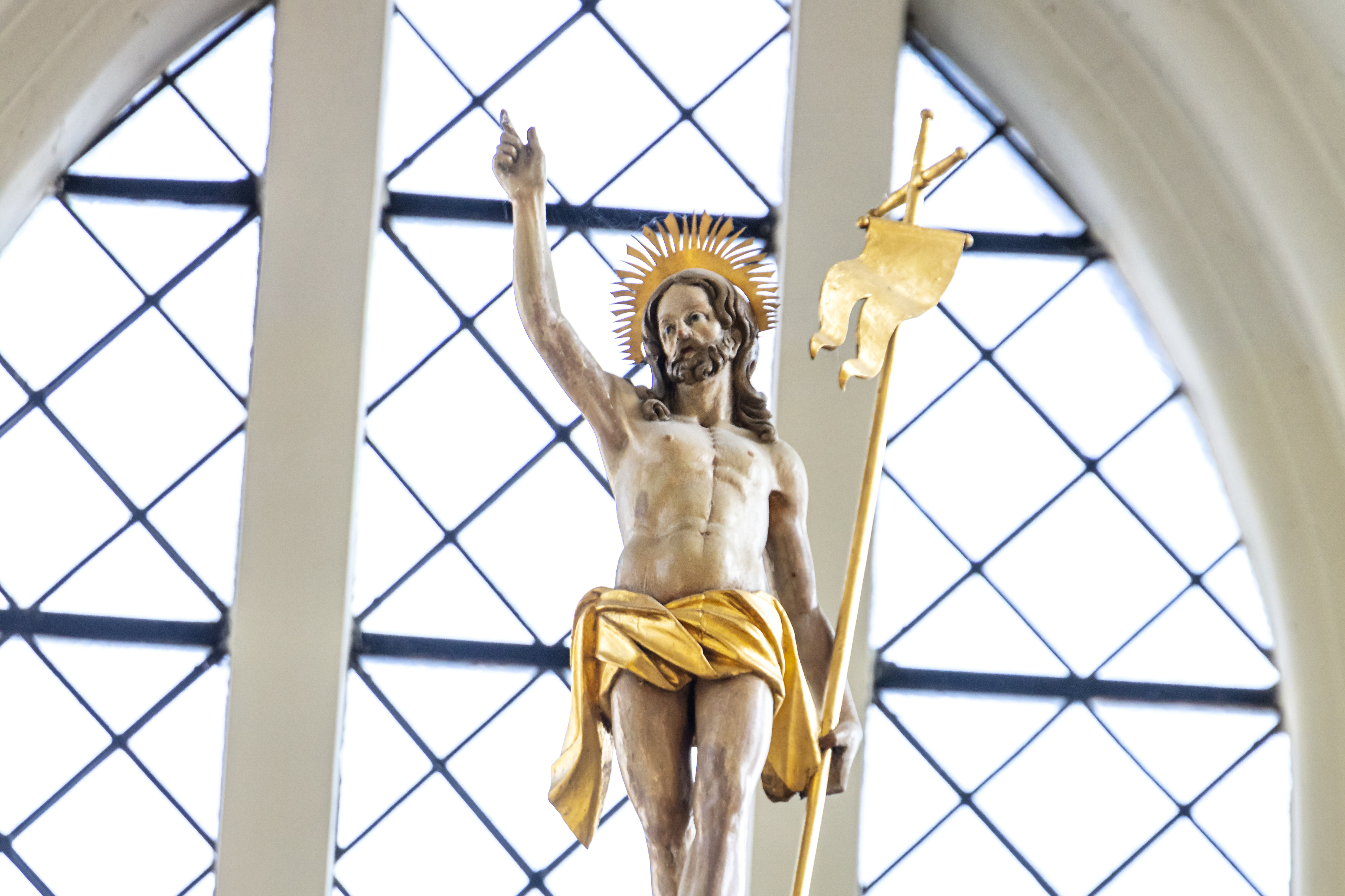 Träfigur som visar en man som höjer fingret mot himlen och i andra handen bär han ett gyllene segerstandar. Han har mörkt långt hår och skägg och är endast iklädd ett gyllene höftskynke.