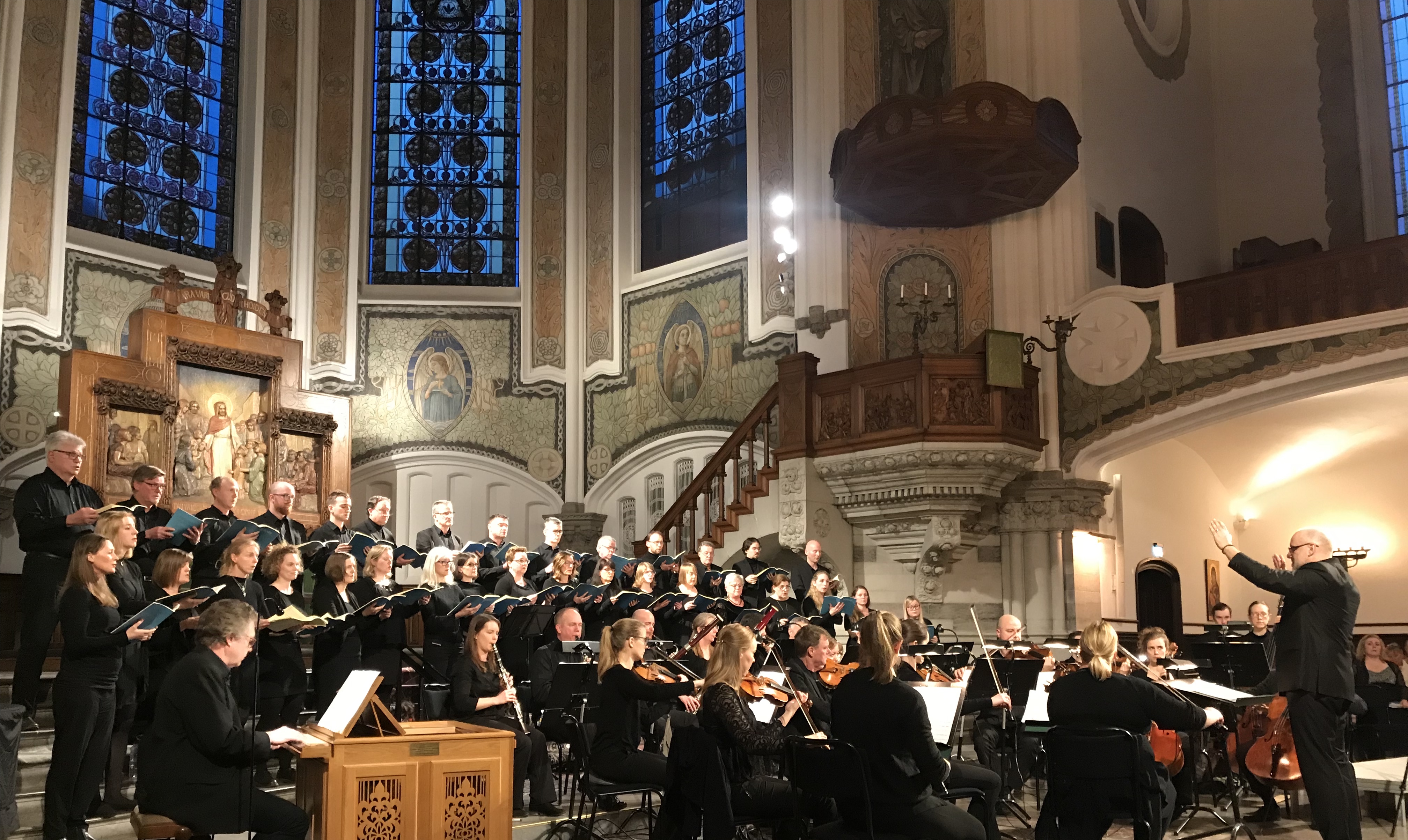 Kör står uppställd framför altaret i kyrka, framför sitter en orkester .